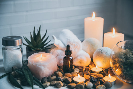 On a table sits a spread of pebbles, several lit candles, a potted succulent, three rolled washcloths, two dryer balls, a small glass globe terrarium, and a shaker of sea salt.